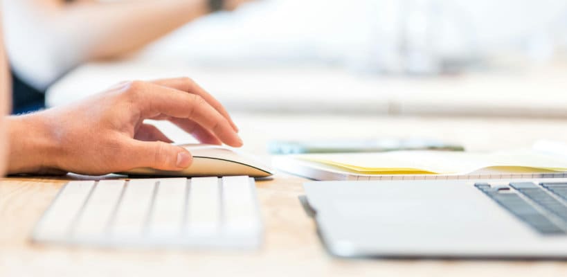 A hand about to click on a mouse button next to a keyboard and a laptop. 